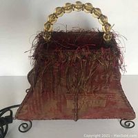 Front angle of purse-shaped accent lamp showing dark red printed fabric shade and bead handle, with attached black power cord coiled to the left.