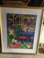 Photo showing framed colorful artwork depicting a kitchen scene with various colored elements including chairs, table, plates, and fruit.