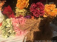 Various large artificial flowers in orange, red, yellow, pink, green, and purple clustered together with some dried ornamental grasses.