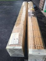 Two rolled bamboo blinds in plastic packaging laid on the ground, showing white wood bases and bamboo slats.