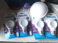 Four Philips LED 10.5W A19 bulbs in original packaging laid flat with one round white push on small space light on a surface.