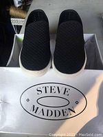Top view of black slip-on sneakers showing perforated mesh upper and white rubber sole inside a Steve Madden box.