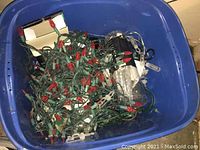 Blue plastic tub containing red bulb Christmas string lights with green wiring, white icicle lights, and other loose seasonal decoration items
