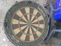 Vintage Nodor Supawire dart board visible showing overall face, numbers and elements, positioned on concrete surface and supported by metal frame.