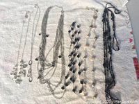 Photo showing all six fashion necklaces laid out on a white textured background with varied styles including silver-tone chains, black beads, and pearl-like beads.
