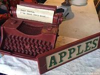 Red Santa-themed typewriter with paper insert saying "Dear Santa, I've been very good..." and wooden APPLES sign side by side on table.