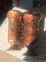 Photo of two tall vases with mosaic tile design laid side by side on a cloth, showing their shape and reflective colorful surfaces under sunlight.