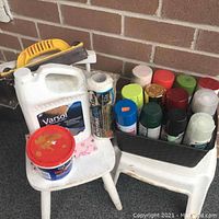 Spray paint cans, paint can, Varsol solvent jug, paper towel roll and scraper on white chairs against brick wall