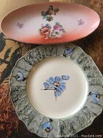 Top view of both plates showing the decorative designs, bird and floral on pink plate; and blue flowers and butterflies with textured rim on round plate.