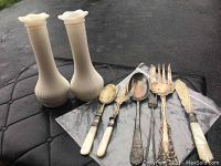 Six pieces of vintage silverware including spoons, serving fork, butter knife, some with mother-of-pearl or bone-like handles, placed next to two white milk glass vases on a black surface outdoors.