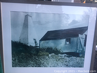 Framed art showing a rural scene with a tall white tapered structure and adjacent building under cloudy sky.