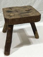 View of the primitive wooden milking stool showing thick square seat with three attached legs and natural wood texture.