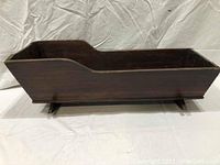 Side view of antique wooden baby cradle showing curved headboard and footboard, dark brown finish with signs of wear.