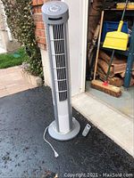 Full view of the Seville Classics upright column fan standing on asphalt near a garage. Gray power cord partially coiled at the base. Remote control placed on the ground.
