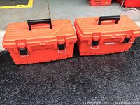 Two orange plastic toolboxes with black handles and metal latches, closed and side by side on ground.