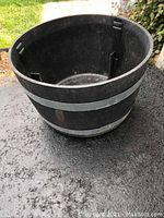 Black simulated oak cask planter pot exterior with metal bands, shown on asphalt surface outdoors.
