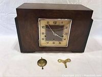 Front view of the clock showing the wooden case, clock face, pendulum and key arranged in front.