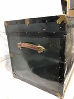 Side view of the black steamer trunk showing brown leather handle and metal corner reinforcements.