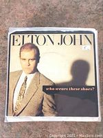 Front-facing image of Elton John 45RPM record sleeve for 'Who Wears These Shoes?' showing the artist in a tan jacket, cream shirt, and patterned tie against a beige background with dark text.