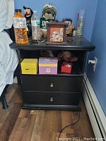 Black wooden nightstand with two drawers, open shelf space, and various objects on top including bottles and plush toys.