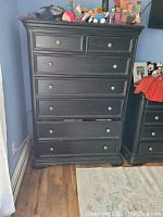Photo of the tall black wood dresser showing front view with seven drawers and metal knobs, placed on wood flooring next to a matching nightstand.