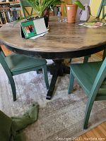 Wooden round table with natural finished top and black painted base set on a rug with two green wooden chairs.