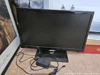 Samsung 25-inch black flat screen TV with power cable on beige carpet, positioned in front of a window and adjacent to a framed picture.