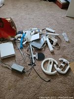 Photo showing complete Nintendo Wii set including multiple Wii Remote controllers, Nunchuck attachments, Wii Wheels, sensor bar, power adapter, AV cable, and the console itself on a carpeted floor.