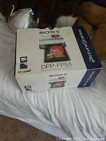 Photo of the Sony DPP-FP55 digital photo printer box placed on a white bedspread with stuffed animals in the background.