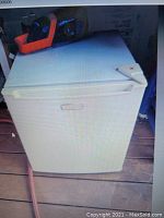 Image showing white mini fridge with top handle and flat top surface, placed on wooden floor.