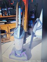 Photo of a purple upright Shark vacuum cleaner with clear dustbin and flexible hose attachment standing upright on a floor next to furniture and sunlight reflecting on the vacuum.