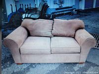 Full frontal view of a beige fabric upholstered loveseat sofa placed outdoors on rough pavement. Shows two seat cushions, two back cushions, rolled armrests, and four wooden block feet.
