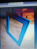 Square wall mirror with blue decorative frame featuring embossed swirl patterns placed on a wooden chest of drawers.