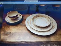 Photo displaying a collection of white dishes and cups with gold leaf design on a wooden surface, showing various sizes of plates and matching teacups and saucers