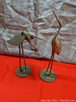 Photo showing two brass stork sculptures, one with the bill directed upward and one with the bill pointing downward. This lot is the upward pointing bill figure only.