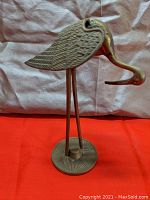 Side view of the brass stork sculpture showing detailed textured wings and downward curved bill on a round base.