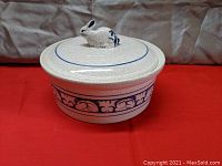Overall front view of cream ceramic casserole dish with rabbit figurine lid handle, blue floral band around base, placed on red fabric background.