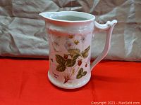 Front side view of white porcelain pitcher decorated with strawberry botanical illustrations, showing handle and spout.