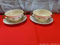 Two Irish Belleek porcelain cream soup bowls with double handles on matching saucers, shamrock pattern, placed side by side on red cloth
