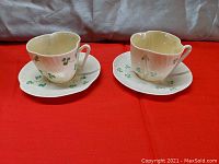 Two Irish Belleek porcelain cups each paired with a matching saucer, all decorated with green shamrock patterns, set on a red surface.