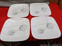 Four white square Corelle plates arranged on a surface showing the scribbled line and spiral design.