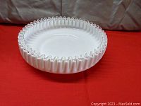 Side view of the white opaque glass shallow bowl with ruffled silver crest edge on a red cloth background.