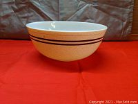 View of large vintage Pyrex mixing bowl exterior showing speckled cream background with two brown horizontal stripes near the rim, set on red fabric background.