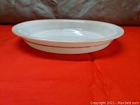 Side view of white Pyrex pie plate with gold band on rim, showing shallow depth and smooth finish, set on red cloth background.
