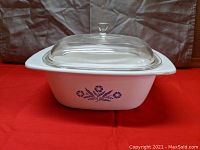 5 quart square white CorningWare Dutch oven with blue cornflower design on side, sitting on red surface with gray background