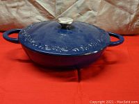 Blue enameled cast iron pot with lid, showing side view with handles and knob lid, resting on red cloth.