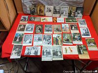 Full view of approximately 30 antique humorous couple scene postcards arranged on a red surface, showing a variety of scenes, handwriting, and some with postage stamps.
