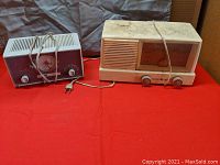 Two vintage radios placed side by side on a red surface, showing General Electric radio on the right in white with cracked case and Philco clock radio on the left with maroon and cream color.