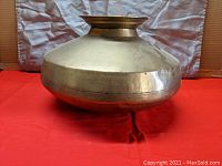 Front view of the large brass vessel showing rounded body and narrow flared neck opening on a red cloth background.