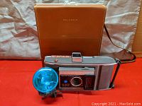 Polaroid Land camera with flash attachment in front of carrying case on red surface